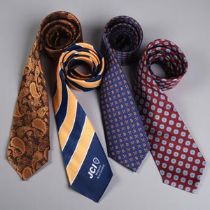 Può includere: Quattro cravatte disposte in fila. La prima è marrone con un motivo paisley dorato. La seconda è a righe blu navy e oro con il testo JCI Malaysia Area Central. La terza è blu navy con un motivo dorato di lettere B e martelli incrociati. La quarta è rossa con un motivo floreale blu e bianco ripetuto.