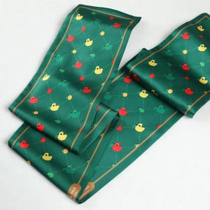 May include: A green satin scarf with a repeating pattern of yellow ducks, red apples, and green pears. The scarf has a gold trim and the text "Fruit Better" is visible on the bottom right corner.