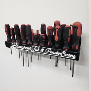 May include: A black metal screwdriver holder with the text "Bluegrass Fab & Powder Coating" on the front. The holder is mounted to a white wall and has 16 screwdrivers with red and black handles.