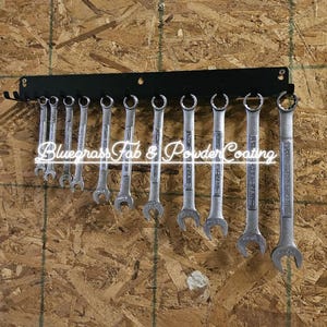 May include: A black metal wrench holder with 12 wrenches hanging from it. The holder is mounted on a wood wall. The text "Bluegrass Fab & Powder Coating" is visible on the holder.