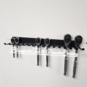 May include: A black metal rack with six hooks for hanging tools.  The rack is mounted on a white wall.  Five silver ratcheting wrenches are hanging on the hooks.  The text "Bluegrass Fab & Powder Coating" is visible on the rack.
