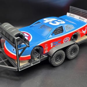 May include: A blue, red, and white race car with the number 43 on a black trailer. The trailer has a rack of tires and a red jack. The car has sponsor logos and the word "Hoosier" on the back.