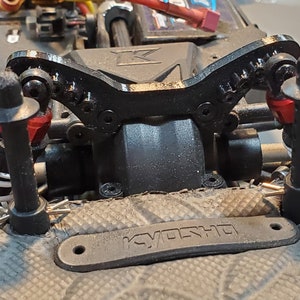 May include: A detailed view of a black RC car chassis component. The image highlights the front suspension, featuring shock absorbers with red detailing, springs, and a black upper support. The word "KYOSHO" is printed on the component.