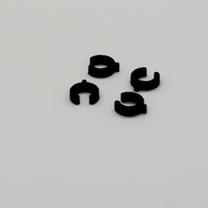 Può includere: Quattro clip per cavi neri, noti anche come fermacavi, sono disposti su una superficie bianca. Questi clip vengono utilizzati per fissare i cavi e i fili in posizione.