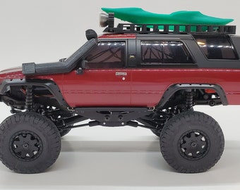 May include: A red toy truck with a black roof rack and a green kayak on top. The truck has large black tires and a black snorkel.
