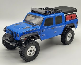 May include: A blue toy Jeep Wrangler with a black roof rack and a black and red storage box on the back. The Jeep has black wheels with silver rims and a black bumper.