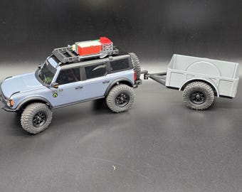 May include: A light blue toy SUV with black wheels and a matching trailer. The SUV has a roof rack with red and white accessories. The trailer is gray and has a simple design, suitable for carrying small items.