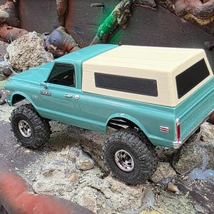 Może przedstawiać: Turkusowy model pick-upa z beżową kabiną kempingową i dużymi czarnymi oponami. Miniaturowa ciężarówka jest przeznaczona do jazdy w terenie, na skalistym tle.