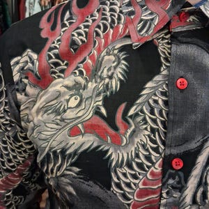 May include: Black button-up shirt with a detailed dragon design in red, gray, and white. The shirt features a classic collar and red buttons. The dragon artwork covers the front and shoulder area of the shirt.