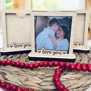 May include: Wooden photo frames with a light wood finish, each featuring a photo of a couple. The frames have a decorative base and the words "love you" with heart accents. A red beaded garland is in the foreground.