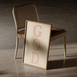 Puede incluir: Una impresión rectangular enmarcada que presenta la palabra GOD en letras grandes de color rojo claro con serifa. Las letras están compuestas por patrones de texto más pequeños y repetitivos. El marco es de madera clara y el fondo es crema.