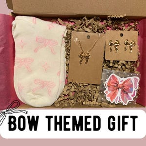 May include: A gift box filled with bow-themed items, including a pair of socks with pink bows, a gold necklace with a bow pendant, a pair of gold bow earrings, and a sticker with a pink bow. The box is labelled "BOW THEMED GIFT".