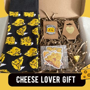 May include: A gift box containing black socks with a yellow cheese pattern, a pin with a smiling cheese wedge and the text CHEESE MAKES EVERYTHING BETTER, a heart-shaped tag, a cheese-shaped keychain, and stickers of cheese wedges on crinkle paper.
