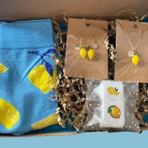 May include: A gift box filled with lemon-themed items. Includes a pair of blue socks with yellow lemon designs, lemon earrings, a lemon pendant necklace, and lemon stickers. The box is lined with blue tissue paper and brown crinkle paper.