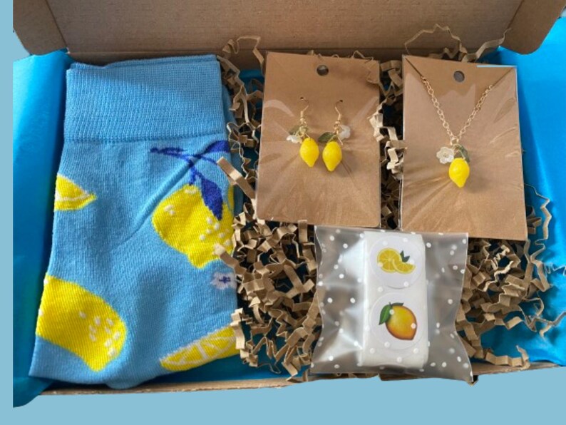 May include: A gift box filled with lemon-themed items. Includes a pair of blue socks with yellow lemon designs, lemon earrings, a lemon pendant necklace, and lemon stickers. The box is lined with blue tissue paper and brown crinkle paper.