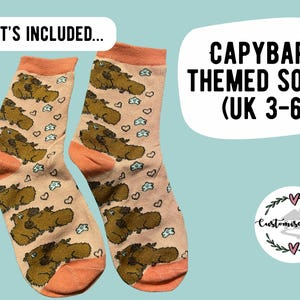 May include: A pair of capybara-themed socks with a light peach background. The socks feature brown capybara illustrations, small blue clouds, and hearts. The cuffs and toes are a darker peach color. Text on the image reads "CAPYBARA THEMED SOCKS (UK 3-6)".