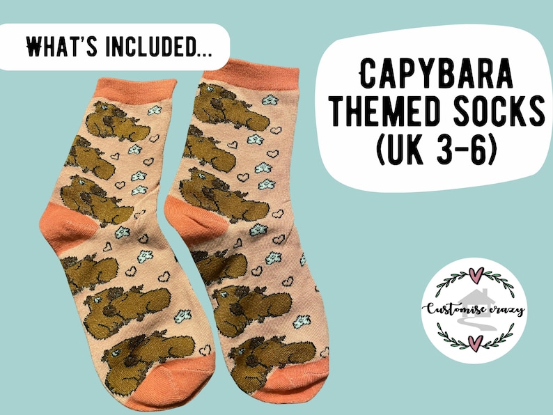 May include: A pair of capybara-themed socks with a light peach background. The socks feature brown capybara illustrations, small blue clouds, and hearts. The cuffs and toes are a darker peach color. Text on the image reads "CAPYBARA THEMED SOCKS (UK 3-6)".