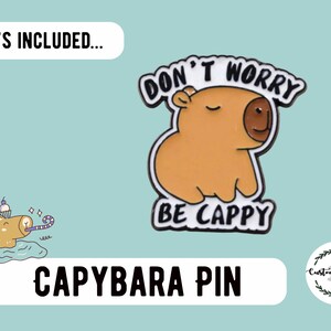 May include: A capybara pin with the text "DON'T WORRY BE CAPPY". The pin features a cartoon capybara in shades of brown and tan with a black outline. The image also includes a small illustration of a capybara with a party hat.