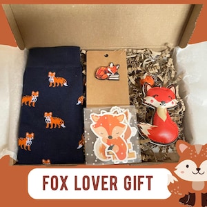 May include: A cardboard gift box containing navy blue socks with a repeating orange fox pattern, a brown card with a fox pin, a clear bag of fox stickers, and a red and white fox keychain, with the text FOX LOVER GIFT at the bottom.