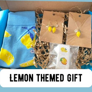 May include: A lemon-themed gift box containing a pair of blue socks with yellow lemon graphics, lemon earrings, a lemon pendant necklace, and lemon stickers. The box is lined with blue tissue paper and brown crinkle paper.