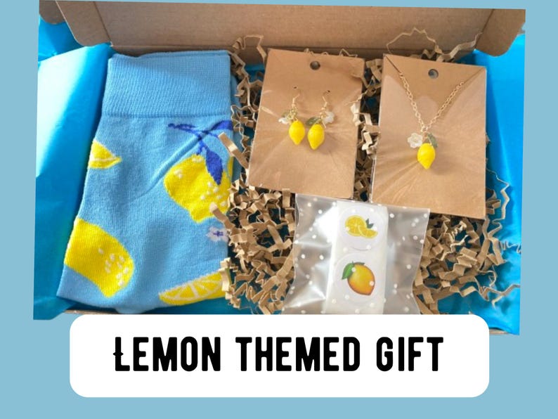 May include: A lemon-themed gift box containing a pair of blue socks with yellow lemon graphics, lemon earrings, a lemon pendant necklace, and lemon stickers. The box is lined with blue tissue paper and brown crinkle paper.