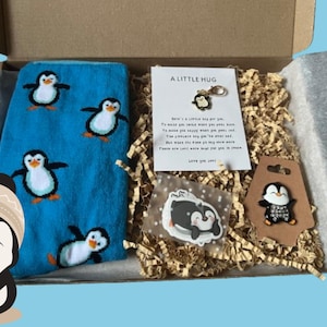 Puede incluir: Un calcet&iacute;n azul y blanco con estampado de ping&uuml;ino, una pegatina de ping&uuml;ino blanco y negro, un amuleto de ping&uuml;ino blanco y negro, una tarjeta marr&oacute;n con un amuleto de ping&uuml;ino blanco y negro y una tarjeta con el texto "A Little Hug" y un mensaje sobre enviar un abrazo.