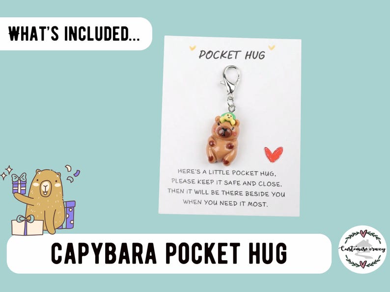 May include: A brown capybara charm with a green hat is attached to a silver keychain. The charm is on a white card that reads "Pocket Hug" and includes a message. The words "Capybara Pocket Hug" are at the bottom.
