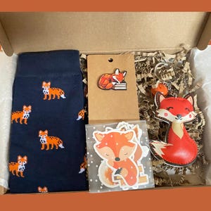 May include: A gift box containing navy blue socks with an orange fox pattern, a brown card with a fox pin, a clear bag with fox stickers, and a fox-shaped keychain. The items are nestled in shredded paper filler within a cardboard box.