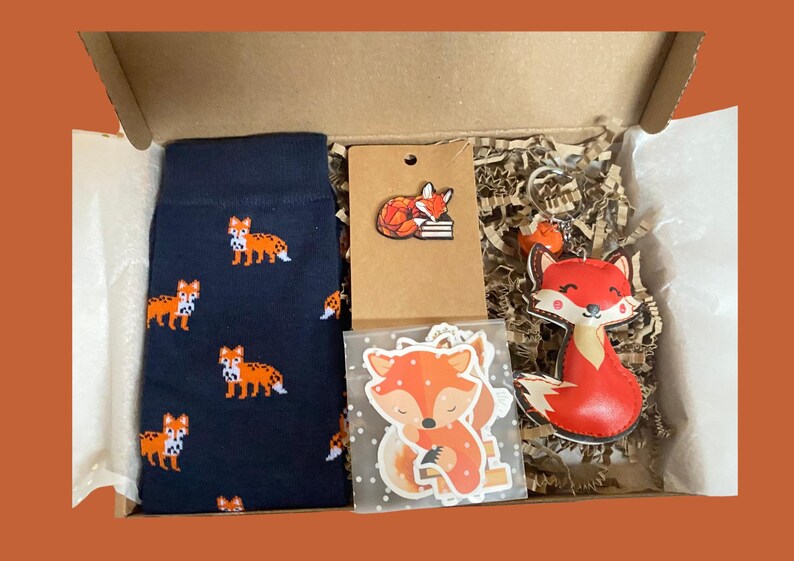 May include: A gift box containing navy blue socks with an orange fox pattern, a brown card with a fox pin, a clear bag with fox stickers, and a fox-shaped keychain. The items are nestled in shredded paper filler within a cardboard box.