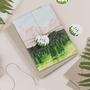 Könnte beinhalten: Ein gefalteter Stoffartikel mit einem Aquarelldruck einer Bergkette und eines dichten grünen Waldes. Ein rundes weißes Etikett, das mit einer Schnur befestigt ist, zeigt den Text J+L über einer kleinen Waldgrafik auf einem grauen Karton.