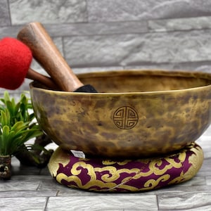 May include: A large, antique-looking brass singing bowl with a dark brown wooden striker and a red felt mallet. The bowl is resting on a purple and gold cushion with a traditional design. The bowl has a circular symbol engraved on the bottom.