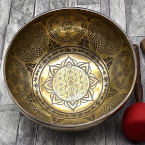 May include: A gold singing bowl with intricate geometric designs, including a flower of life pattern. The bowl is accompanied by two wooden strikers, one with a black rubber tip and the other with a red felt tip.