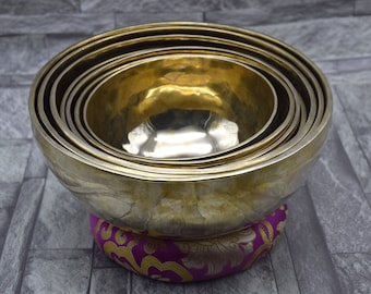 May include: A set of seven nested singing bowls, made of brass, with a hammered finish. The bowls are arranged in a stack, with the largest bowl at the bottom and the smallest bowl at the top. The bowls are resting on a purple and gold cushion.