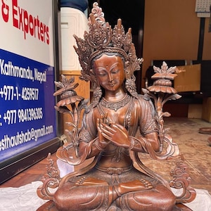 Może przedstawiać: Miedziana statua siedzącego bóstwa z koroną i bogatymi ozdobami. Statua trzyma ręce złożone w geście modlitwy. Statua siedzi na podstawie w kształcie lotosu.