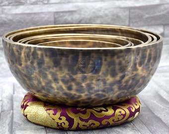 May include: A set of seven nested singing bowls, made of brass, with a hammered finish. The bowls are arranged in a stack, with the largest bowl at the bottom and the smallest bowl at the top. The bowls are resting on a purple and gold cushion with a floral pattern. The largest bowl has a symbol engraved on the inside.