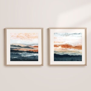 May include: Two framed abstract landscape paintings with a muted color palette of blue, brown, and white. The paintings are in wooden frames and are hung on a white wall.