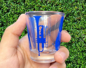 May include: A clear glass shot glass with a blue silhouette of musical instruments, including a trumpet, saxophone, and a bass clef.