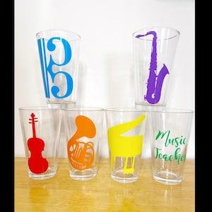 May include: Six clear glass cups with different musical instrument designs. The cups are arranged in a row on a wooden surface. The designs include a treble clef, a saxophone, a violin, a french horn, a piano, and the words "Music Teacher".