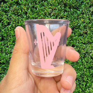 Puede incluir: Un vaso de chupito de vidrio transparente con un diseño de arpa rosa. El vaso de chupito se está sosteniendo en una mano.