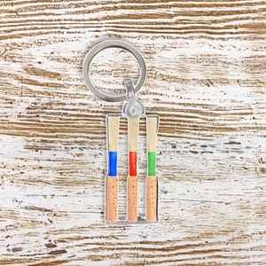 May include: A silver keychain with three wooden oboe reeds. The reeds are painted with blue, red, and green accents.