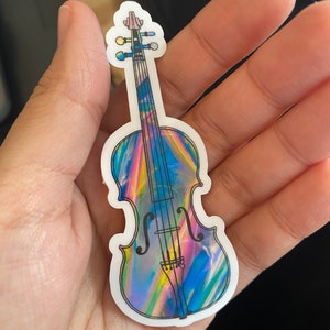 Puede incluir: Una pegatina hologr&aacute;fica de un viol&iacute;n con un acabado iridiscente arco&iacute;ris. El viol&iacute;n es negro y blanco con un dise&ntilde;o detallado.