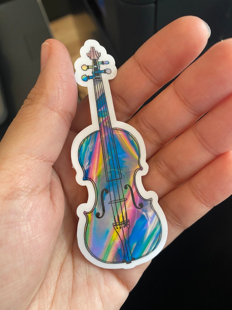 Puede incluir: Una pegatina hologr&aacute;fica de un viol&iacute;n con un acabado iridiscente arco&iacute;ris. El viol&iacute;n es negro y blanco con un dise&ntilde;o detallado.