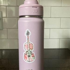 Puede incluir: Una botella de agua de color morado claro con una pegatina de viol&iacute;n con estampado floral en el lateral.