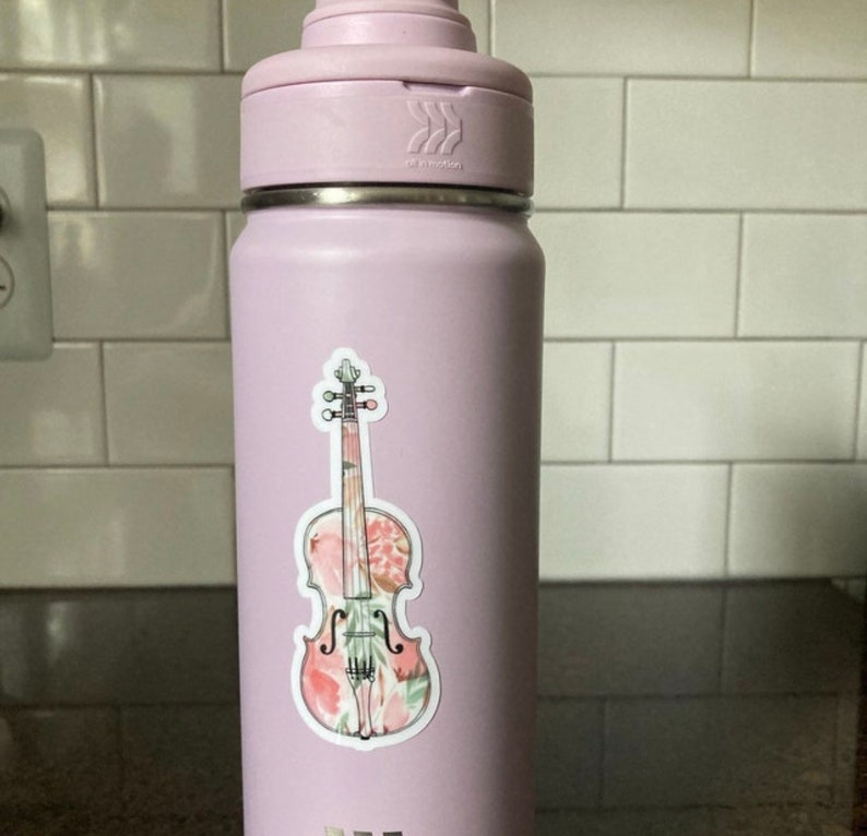 Puede incluir: Una botella de agua de color morado claro con una pegatina de viol&iacute;n con estampado floral en el lateral.