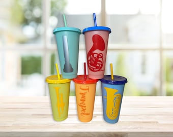 May include: Five colorful reusable tumblers with different musical instruments printed on them. The tumblers are arranged in a pyramid shape with the top tumbler being pink with a French horn, the next two tumblers are blue and green, and the bottom two tumblers are yellow and orange with a trumpet and a tuba.