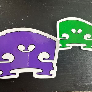 May include: Two bass bridge stickers, one purple and one green, with a white outline. The stickers are shaped like a bass bridge and have a stylized design of the bridge's f-holes.