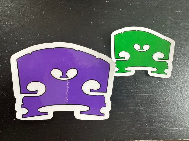 May include: Two bass bridge stickers, one purple and one green, with a white outline. The stickers are shaped like a bass bridge and have a stylized design of the bridge's f-holes.