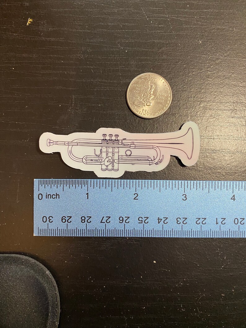 May include: A white and purple outline sticker of a trumpet with a silver US quarter for scale. The sticker is approximately 3 inches long.