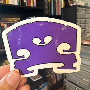 May include: A purple and white sticker shaped like a bass guitar bridge. The sticker is slightly worn.