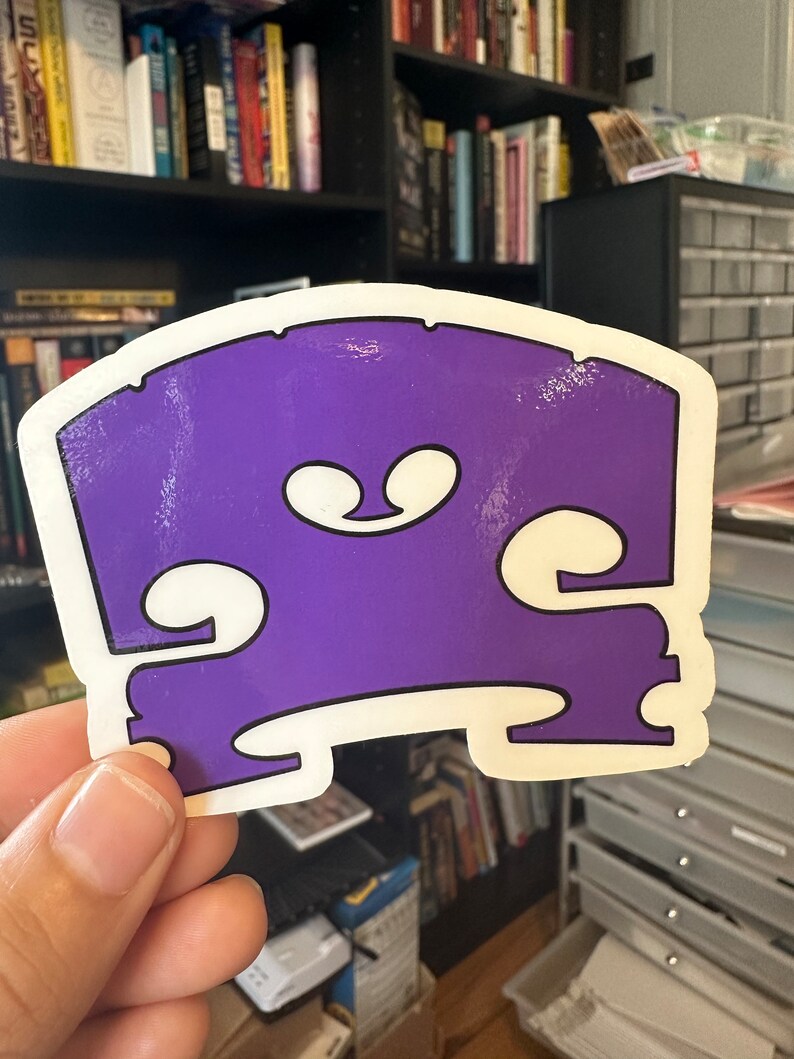 May include: A purple and white sticker shaped like a bass guitar bridge. The sticker is slightly worn.